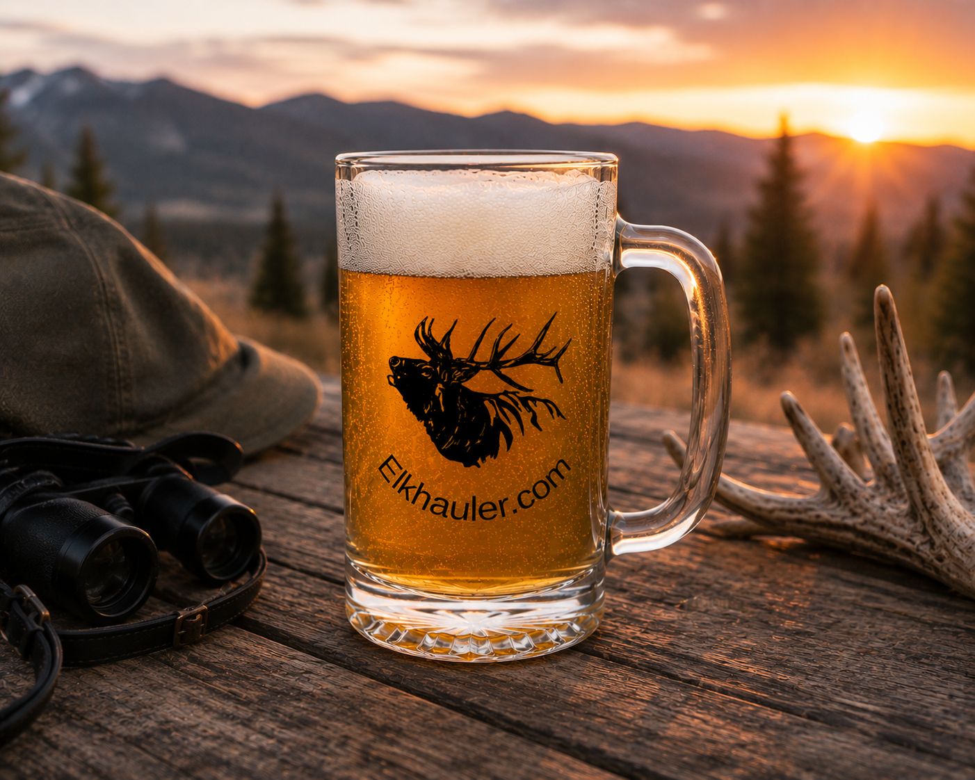 Glass of beer with elk logo and 'Elkhauler.com' text on a wooden surface with sunset and mountains in the background.