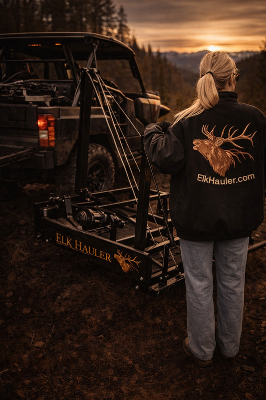 Person wearing a jacket with 'ElkHauler.com' logo standing next to an Elk Hauler vehicle at sunset.